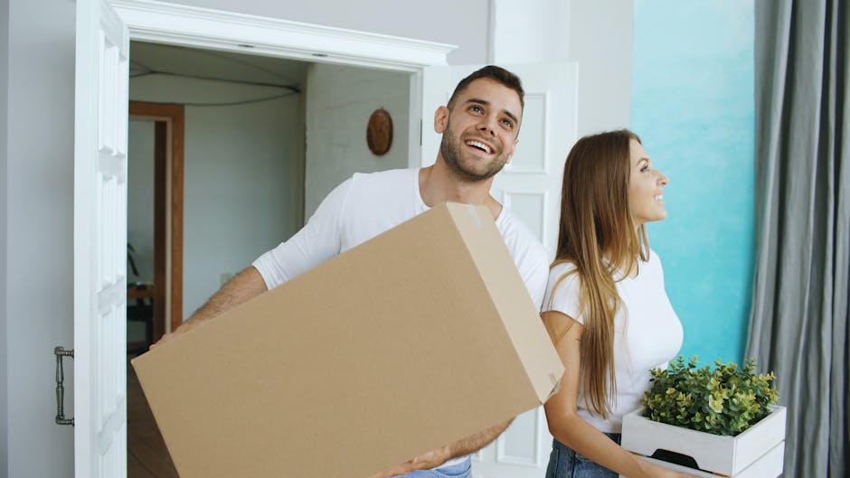 Two young women with long, dark hair smiling while taking a selfie inside a property surrounded by stacked cardboard boxes and packing materials, including plastic-wrapped items and bubble wrap, indic
